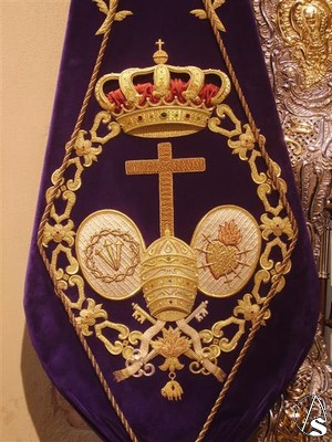  El escudo de la hermandad lo forma la cruz de animas y a sus lados un ovalo, en uno de ellos la corona de espinas y los tres clavos y en el otro un corazón con los siete puñales, rodea todo ello el toisón de oro y remata la corona real. 